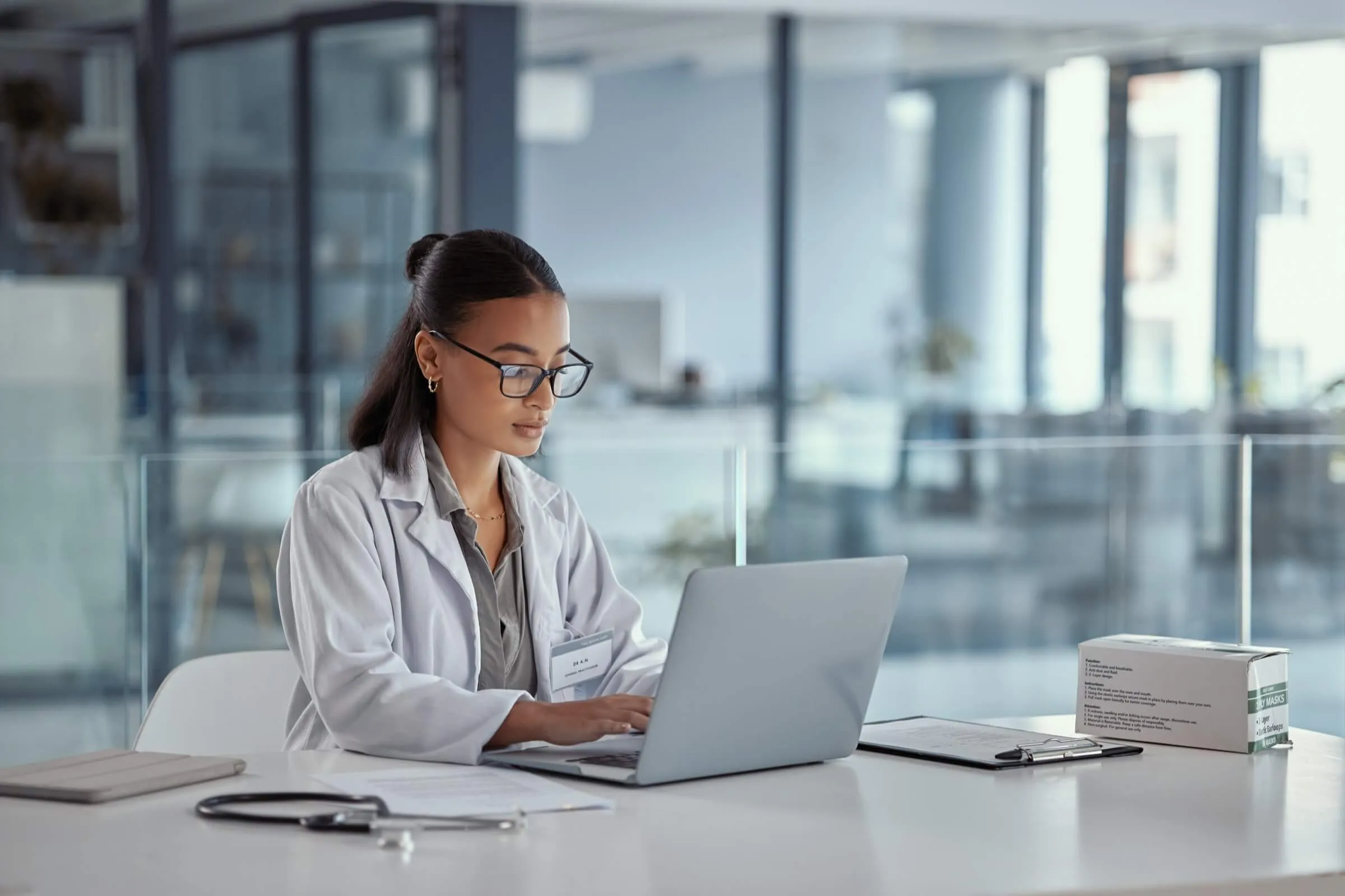 Healthcare professional in white coat working on laptop in modern medical office setting.